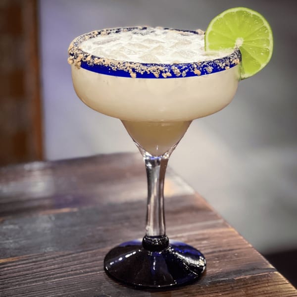 A Tacos El Tio margarita with a salted rim and lime wedge, on a wooden table.