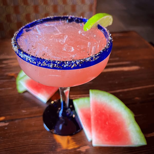 Pink watermelon margarita with a blue rim, salt, lime, and watermelon slices on a wooden table.