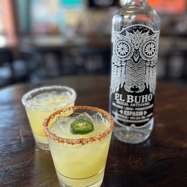 Two margaritas, one garnished with a jalapeño slice, next to a bottle of El Buho mezcal.