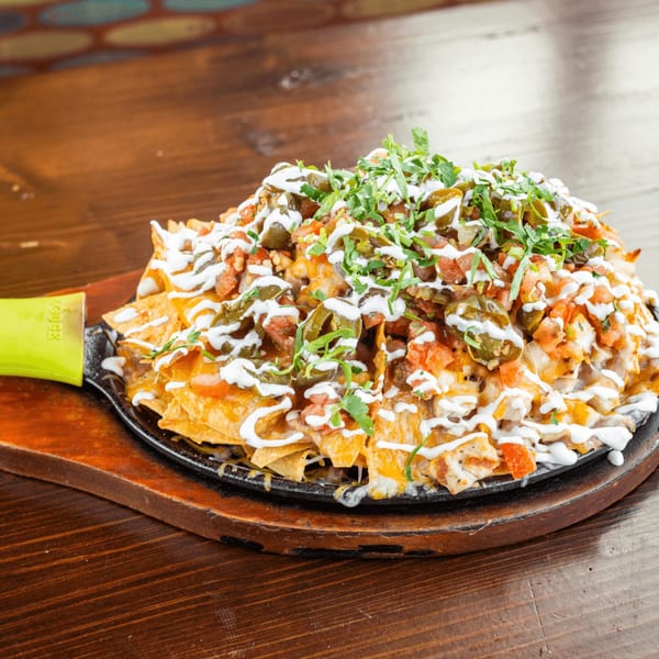Tacos El Tio's loaded nachos with cheese, jalapeños, tomatoes, and sour cream on a sizzling platter.