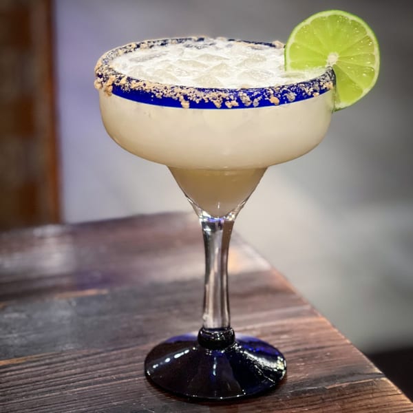 A Tacos El Tio margarita with a blue rim, salt, and a lime wedge on a wooden table.