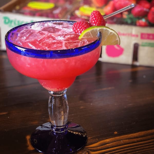 Raspberry margarita in a blue-rimmed glass, garnished with lime and raspberries, on a wooden table.