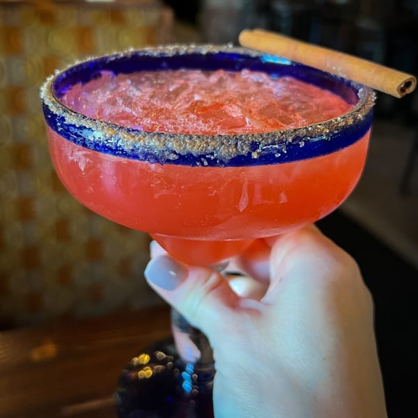 A hand holds a vibrant red margarita with a blue rim, salt, and a cinnamon stick.