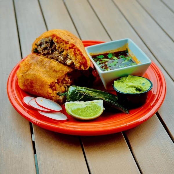 Two deep-fried burritos with rice and meat, served with salsa, guacamole, radish, lime, and a pepper.