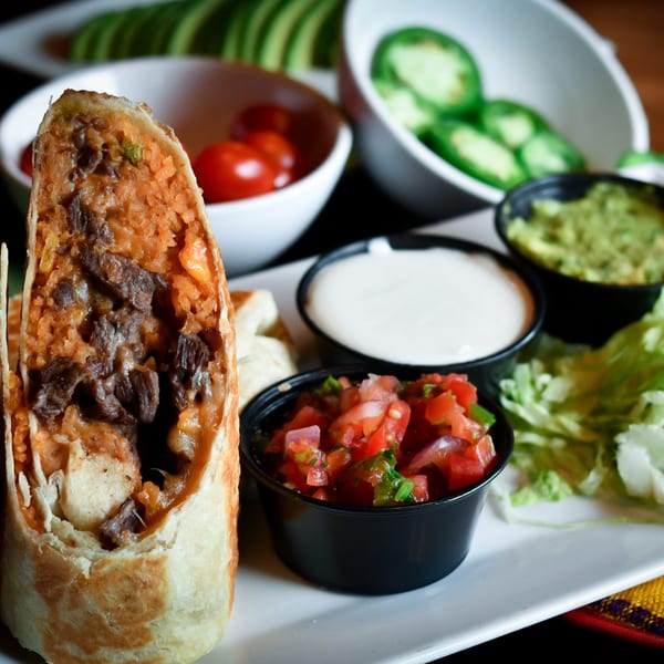 A Tacos El Tio burrito, cut in half, with sides of salsa, guacamole, sour cream, and lettuce.