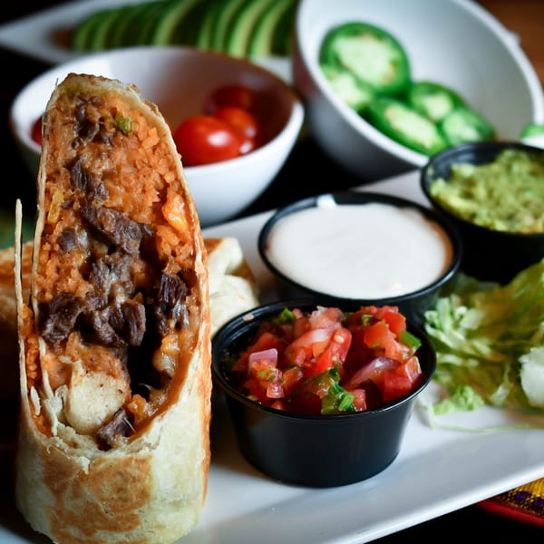 A Tacos El Tio burrito, cut in half, with sides of salsa, guacamole, sour cream, and jalapeños.