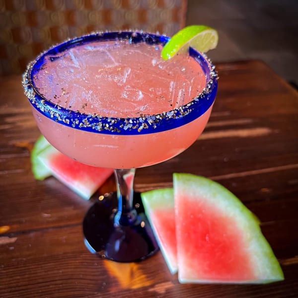 Watermelon margarita with a blue rim and lime, surrounded by watermelon slices on a wooden table.