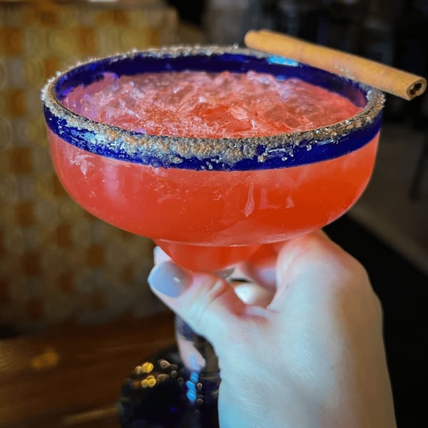 A hand holds a vibrant red margarita with a blue rim, sugar, and a cinnamon stick.