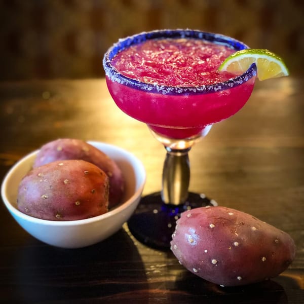 A vibrant pink prickly pear margarita with a lime wedge, next to a bowl of prickly pears.