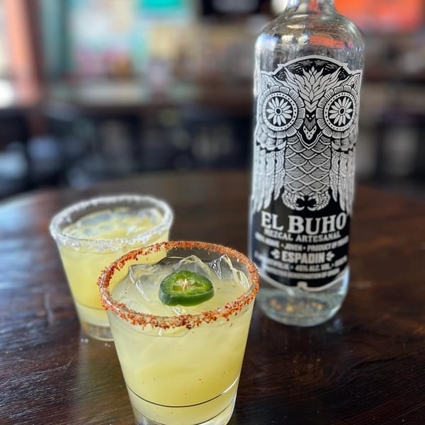 Two margaritas, one with a chili-lime rim and jalapeño, next to an El Buho mezcal bottle.